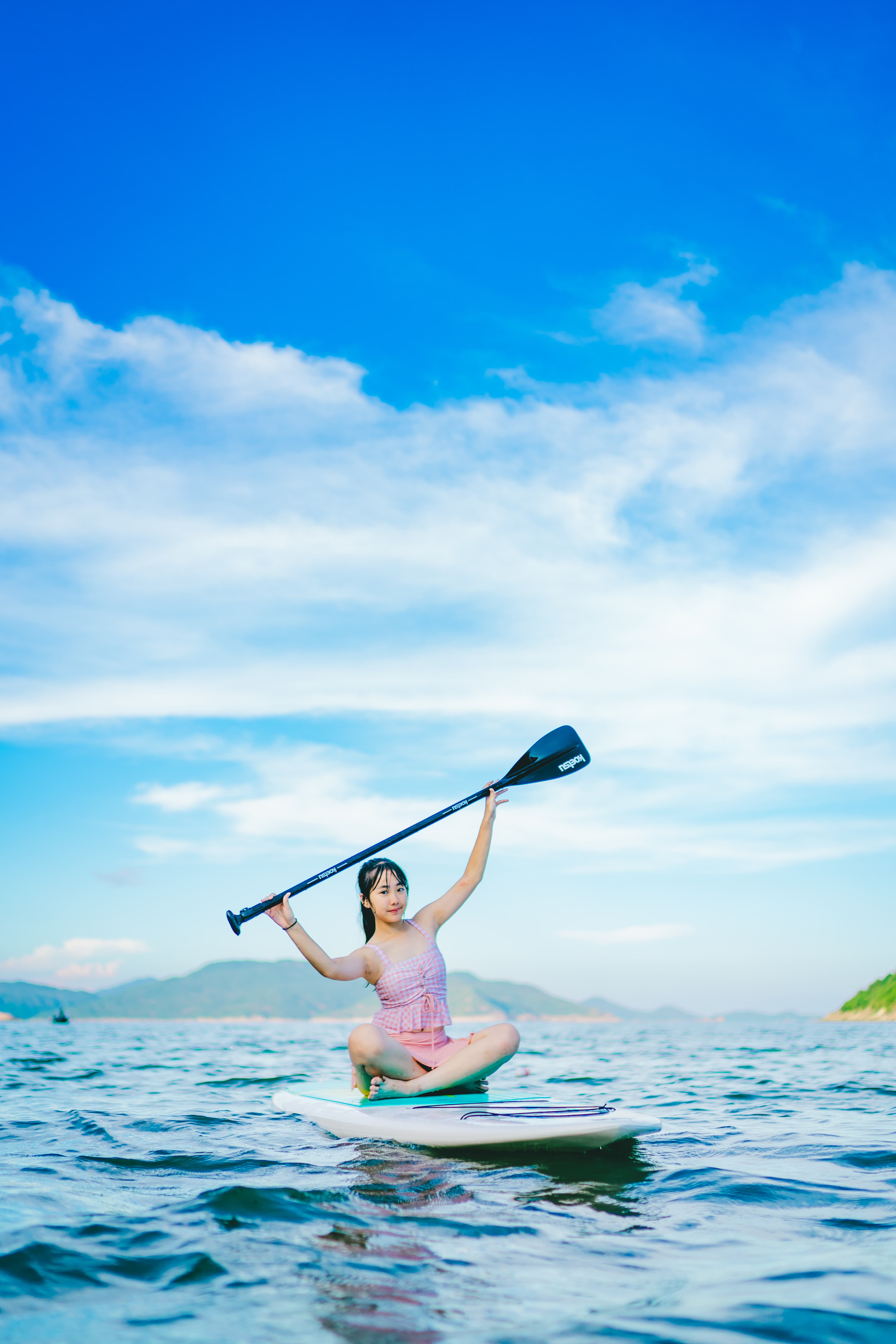 一名女孩穿着粉色泳衣坐在海上桨板上,双手高举船桨,背景是晴朗蓝天。